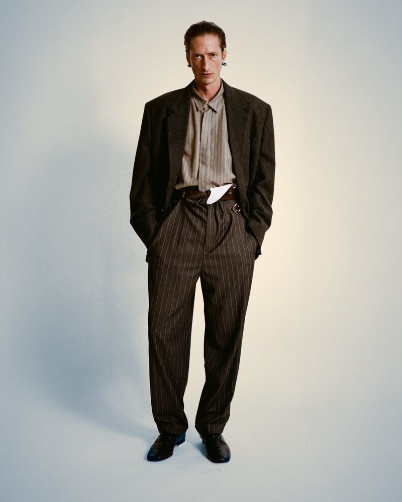 Full-length studio shot of an adult male fashion model with auburn hair wearing a dark suit, pinstripe pants, and a collared shirt.
