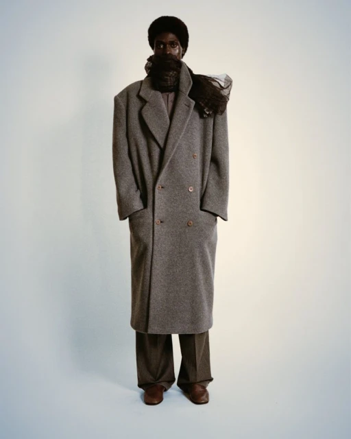 Full-length studio shot of a Black male model wearing a long grey coat and brown trousers, posing against a neutral background.