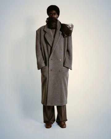 Full-length studio shot of a Black male model wearing a long grey coat and brown trousers, posing against a neutral background.