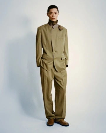 An Asian man in a tan suit stands in a photo studio with a neutral expression, showcasing high-fashion menswear attire and polished leather shoes.