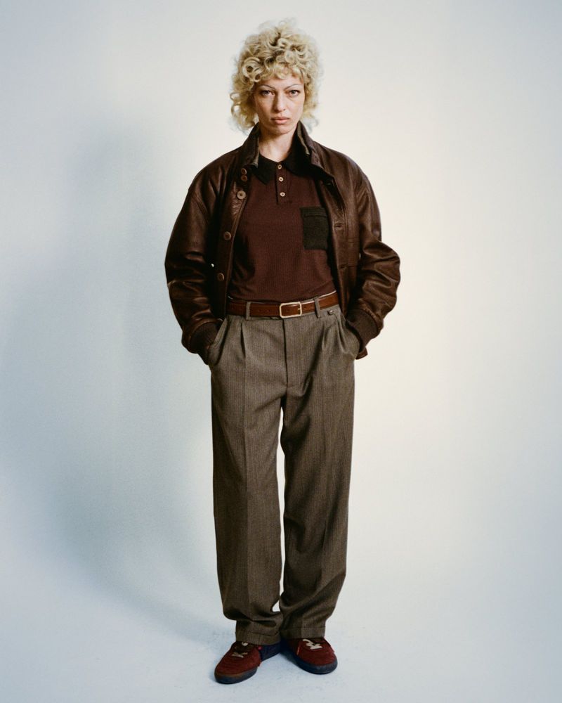 Full-length studio portrait of a woman with blonde curly hair wearing a brown leather jacket, brown polo shirt, grey slacks, and brown shoes.