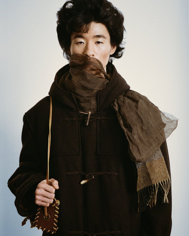 Portrait of an adult male model wearing a dark brown hooded coat, tan scarf, and carrying a small brown pouch against a neutral backdrop.