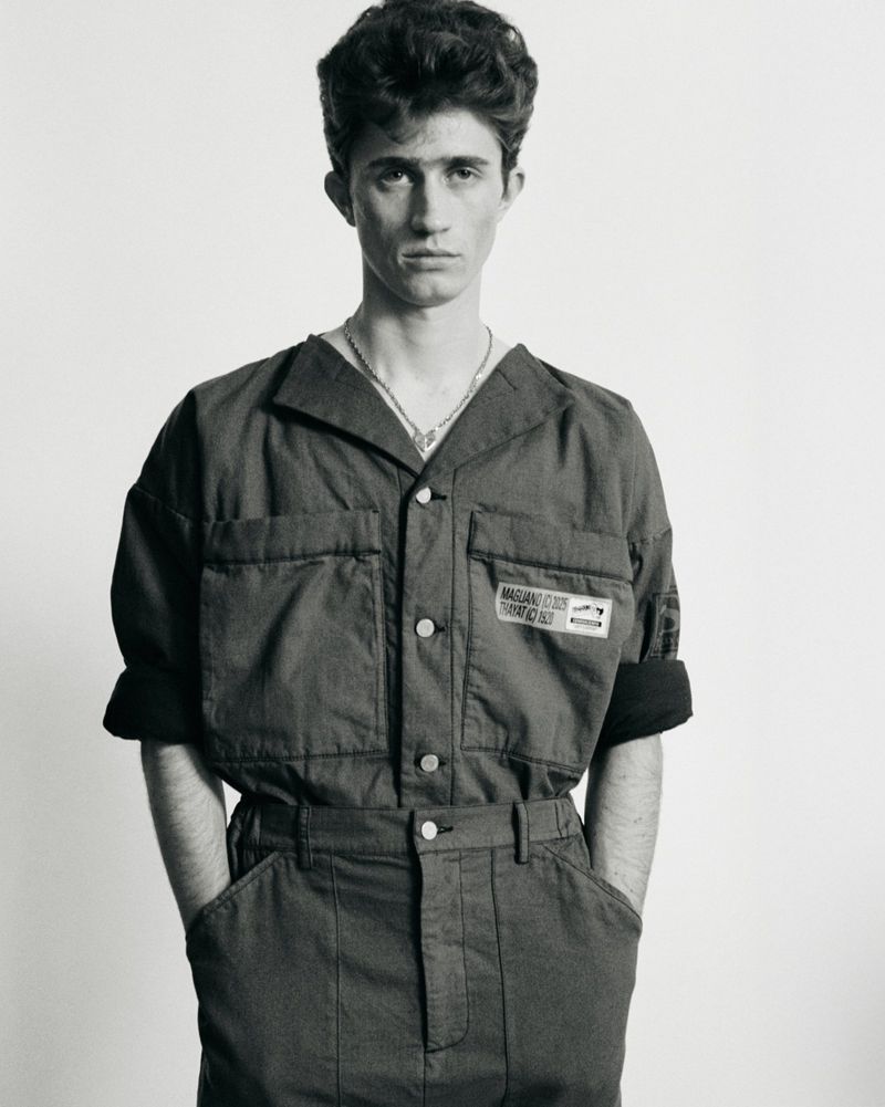 Monochrome portrait of a young adult male in an oversized blue jumpsuit against a white backdrop, captured in a studio setting with soft lighting.