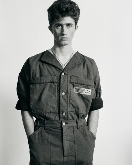 Monochrome portrait of a young adult male in an oversized blue jumpsuit against a white backdrop, captured in a studio setting with soft lighting.