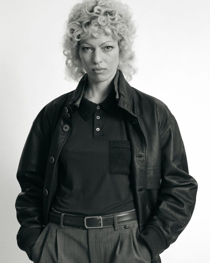 Monochrome portrait of an adult woman with curly blond hair, wearing a dark leather jacket and a dark polo shirt, in a studio.
