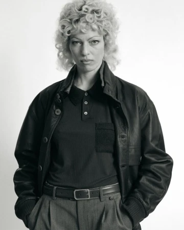 Monochrome portrait of an adult woman with curly blond hair, wearing a dark leather jacket and a dark polo shirt, in a studio.