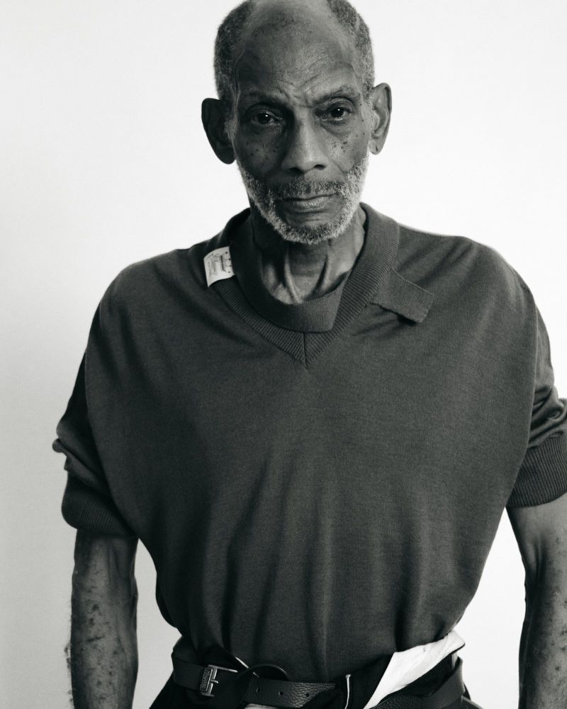 Monochrome portrait of an elderly Black man in a v-neck shirt and belt, set against a bright white backdrop in a studio environment.