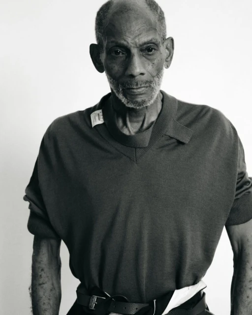 Monochrome portrait of an elderly Black man in a v-neck shirt and belt, set against a bright white backdrop in a studio environment.