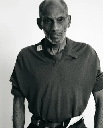Monochrome portrait of an elderly Black man in a v-neck shirt and belt, set against a bright white backdrop in a studio environment.