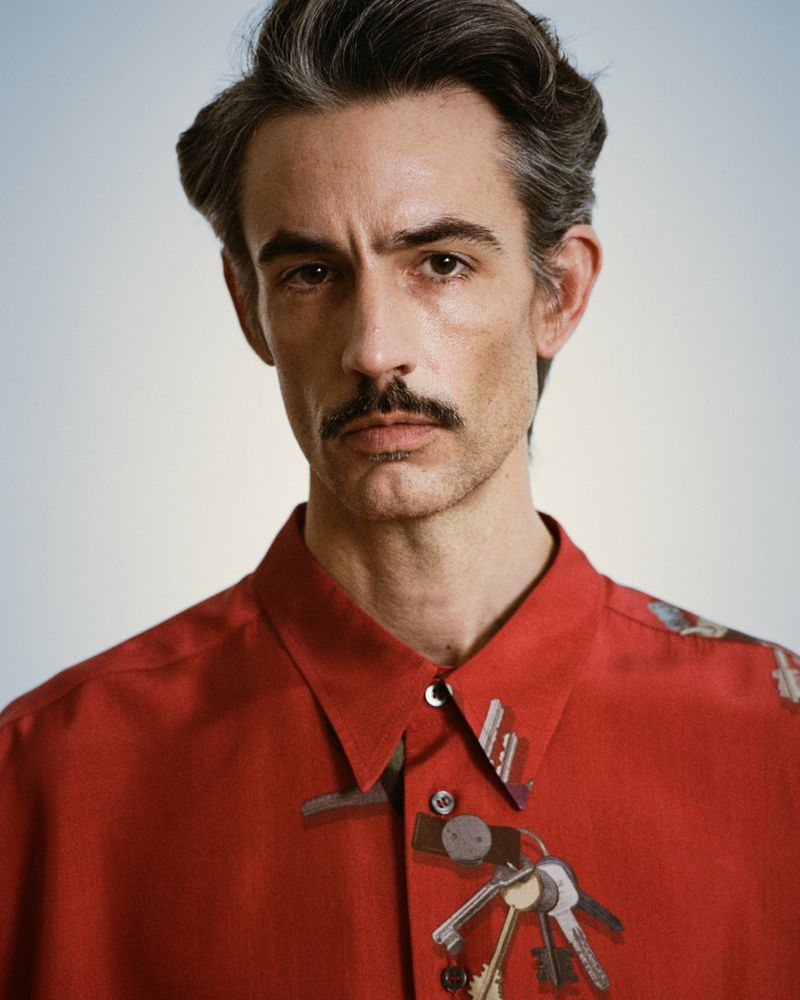 Portrait of a male with a mustache wearing a red shirt and a collection of keys in an eye-level studio shot, captured with photography.
