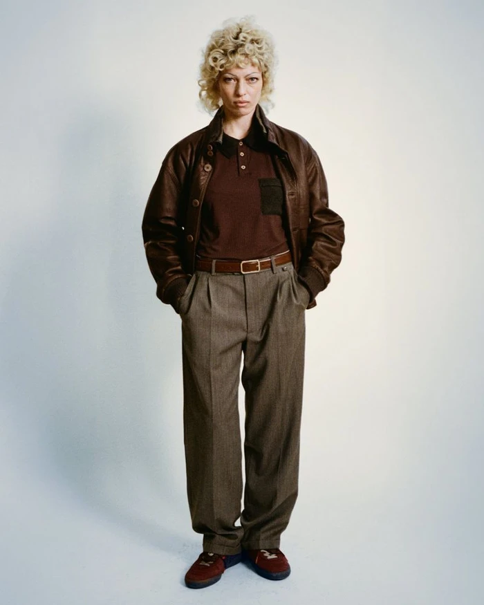 Model with blonde curly hair wearing a brown leather jacket and pleated trousers against a neutral background