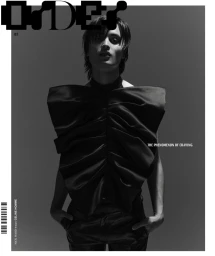 Monochrome portrait of a male model wearing a sculptural black top with hands in pockets against a grey studio backdrop.