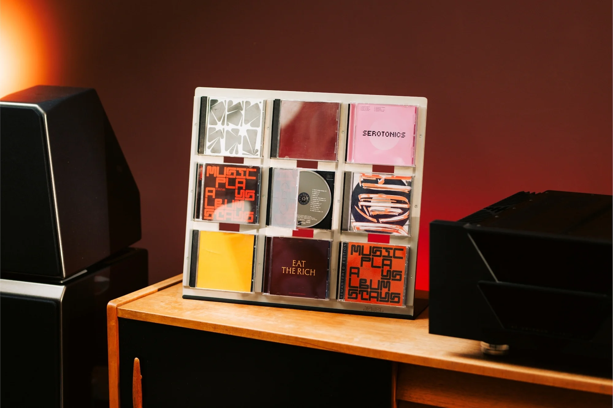 Compact disc collection in a white rack on a wooden table next to a black audio speaker against a solid red-brown background.