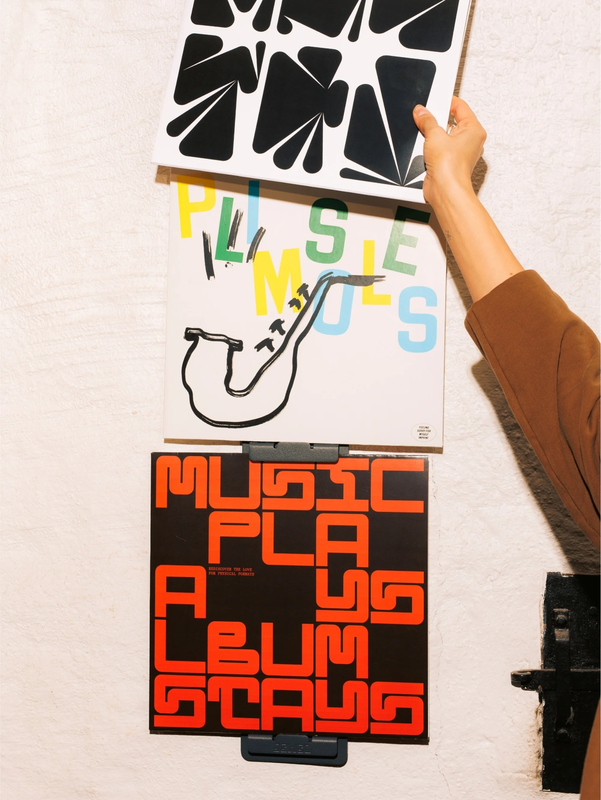 Three album covers are displayed on a wall. A hand reaches to adjust the top cover, which has a black and white geometric pattern.