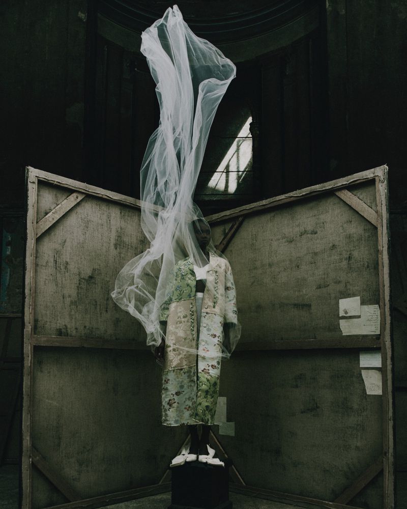 A person in a patterned coat stands on a pedestal in an old room with a veil flowing above their head.