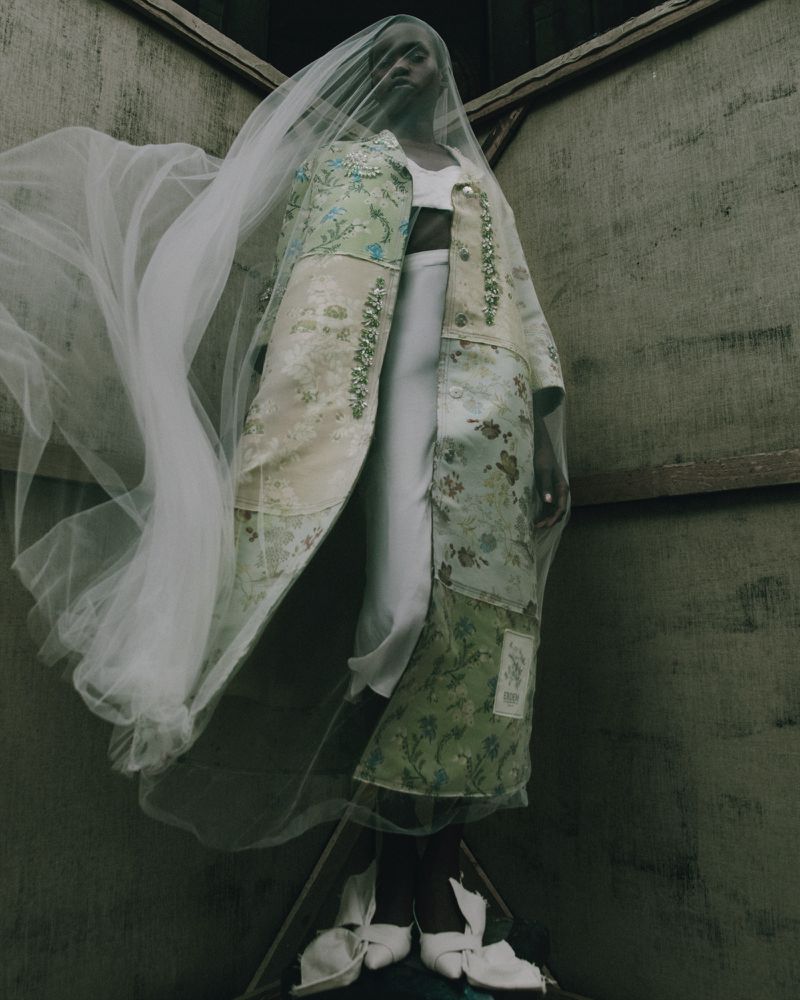 Model wearing a floral coat and veil standing in a corner. Fashion shot with indoor setting, neutral color palette, and high angle.