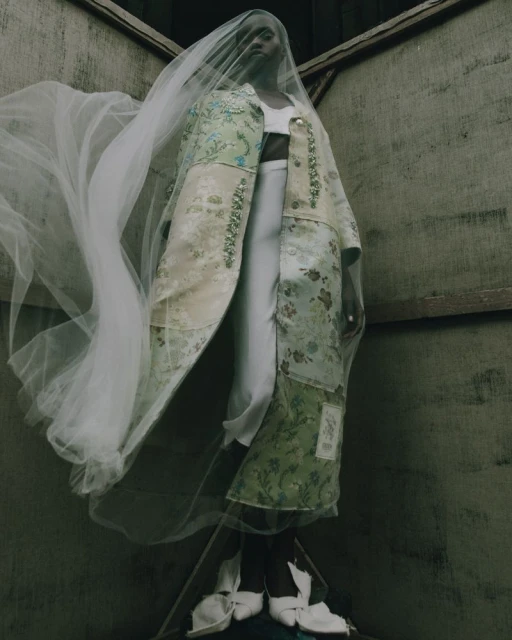 Model wearing a floral coat and veil standing in a corner. Fashion shot with indoor setting, neutral color palette, and high angle.