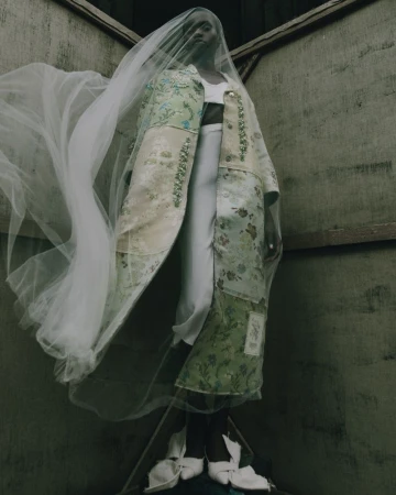 Model wearing a floral coat and veil standing in a corner. Fashion shot with indoor setting, neutral color palette, and high angle.