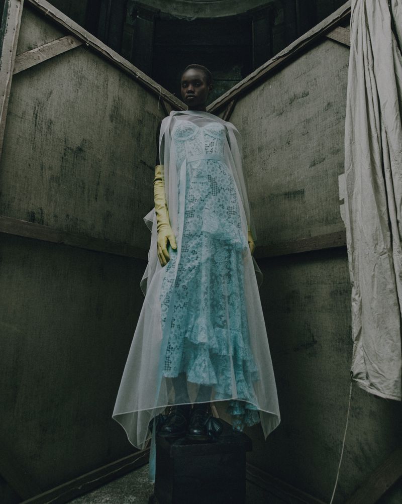 Woman in a light blue lace dress with a sheer cape and yellow gloves, stands on a pedestal against an abstract angular background.