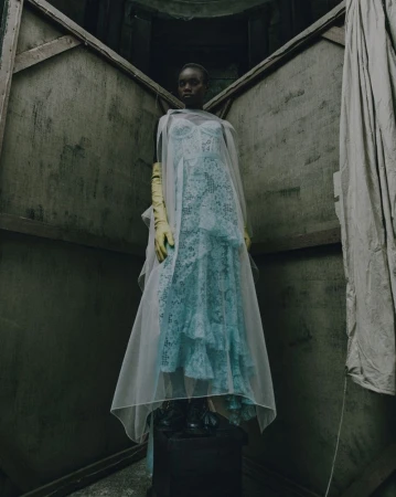 Woman in a light blue lace dress with a sheer cape and yellow gloves, stands on a pedestal against an abstract angular background.