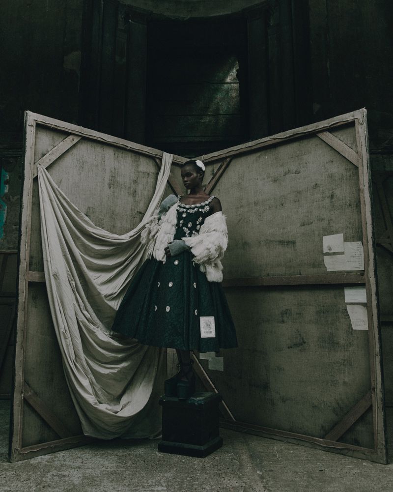 Fashion shot of an African woman wearing a vintage dress with white flowers and a fur stole standing on a pedestal in an indoor set.