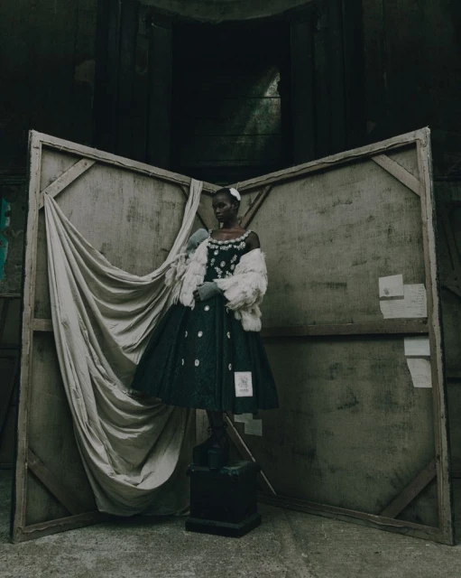 Fashion shot of an African woman wearing a vintage dress with white flowers and a fur stole standing on a pedestal in an indoor set.