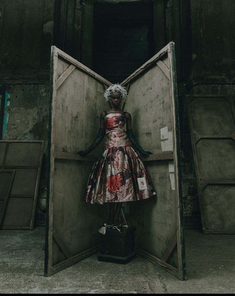Model wearing a floral print dress and dark gloves standing framed by large wooden panels in an atmospheric interior.