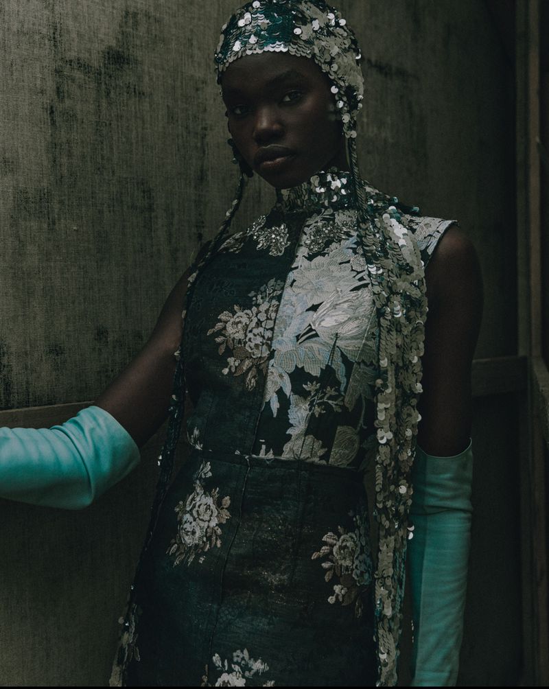 Elegant dark-skinned woman in floral high-neck dress and sequined headpiece, posing indoors against mottled textured wall.