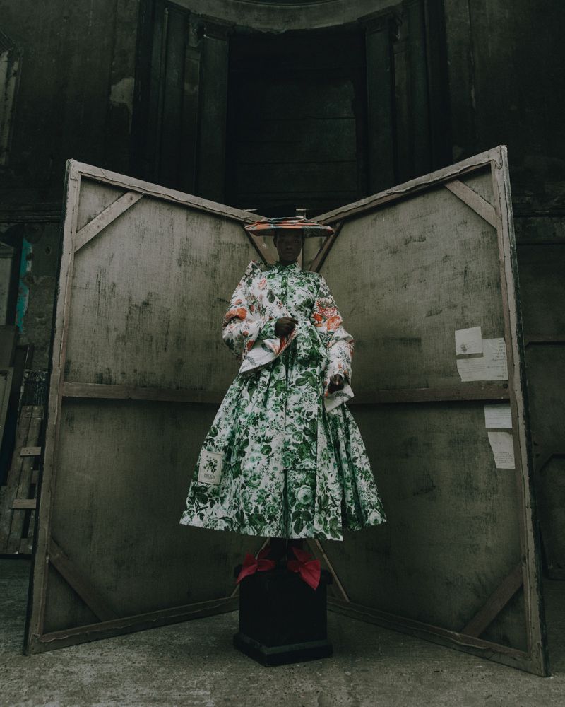 Adult female fashion model in a floral dress, wearing a unique hat, posing indoors in front of a weathered wooden structure and black pedestal.