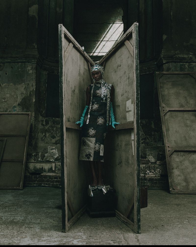 A Black fashion model poses inside a deconstructed art easel in an aged interior space. The model wears a dress, gloves, and head covering.