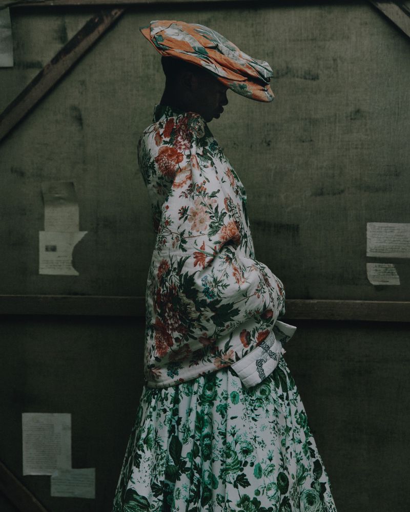 Model wearing a patterned floral jacket and matching skirt with a sculpted headpiece against a dark green textured studio background.