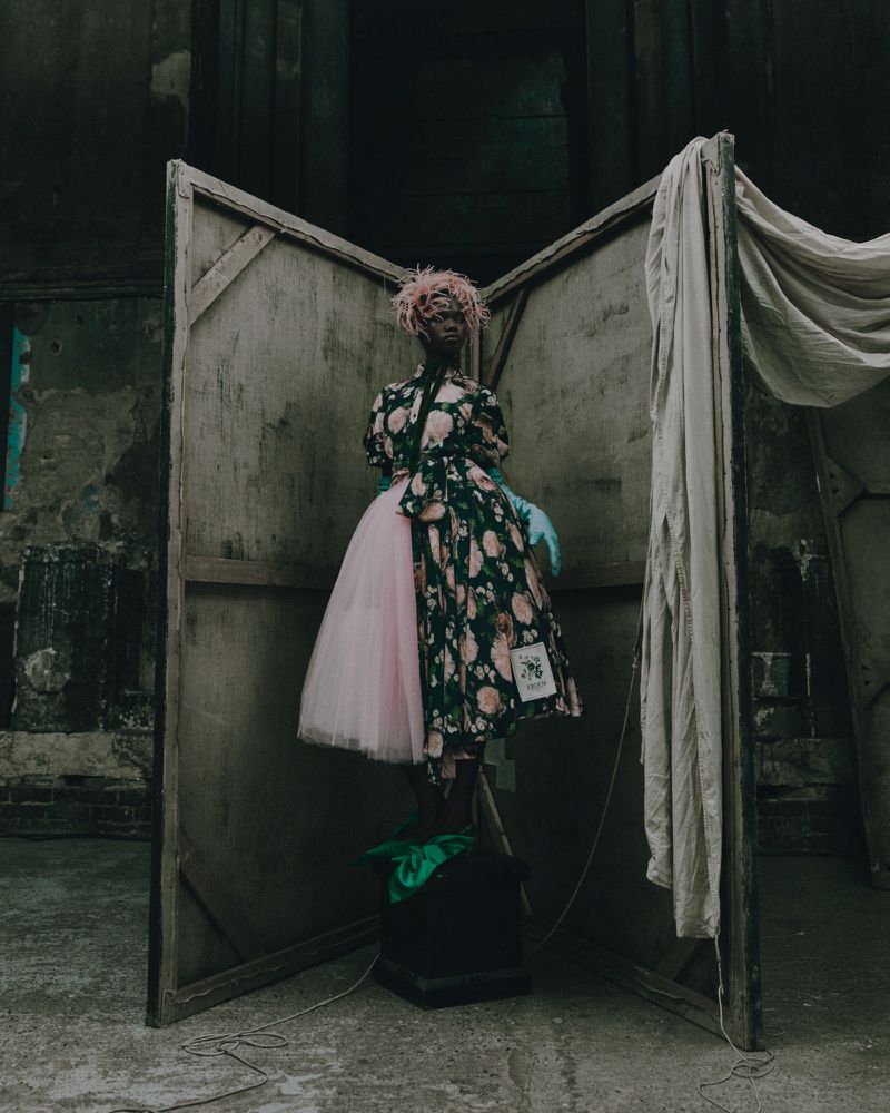 A person in a floral dress and pink tutu stands in front of a weathered, wooden backdrop inside a derelict building.