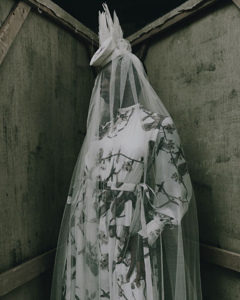 Woman in a patterned dress and white veil stands in an indoor space with canvas walls, creating a unique and artistic fashion statement.
