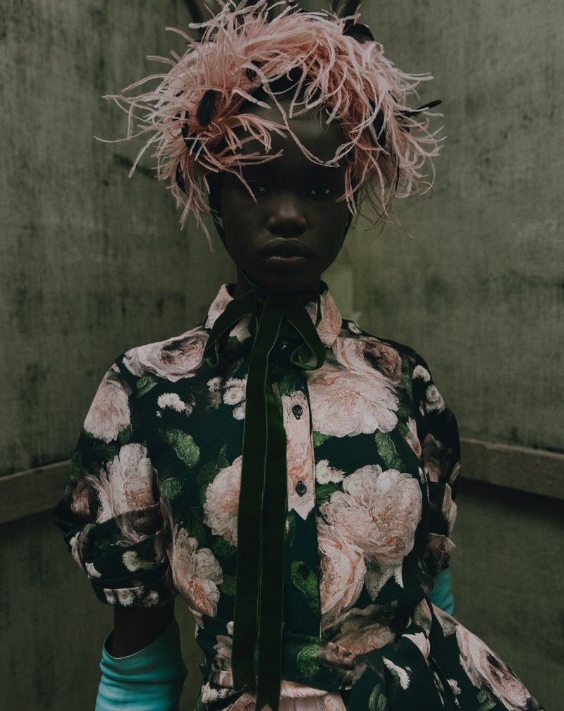 Fashionable woman with dark skin poses in floral dress, adorned with a feathered hat and dark green velvet ribbon tie, against a grey wall.
