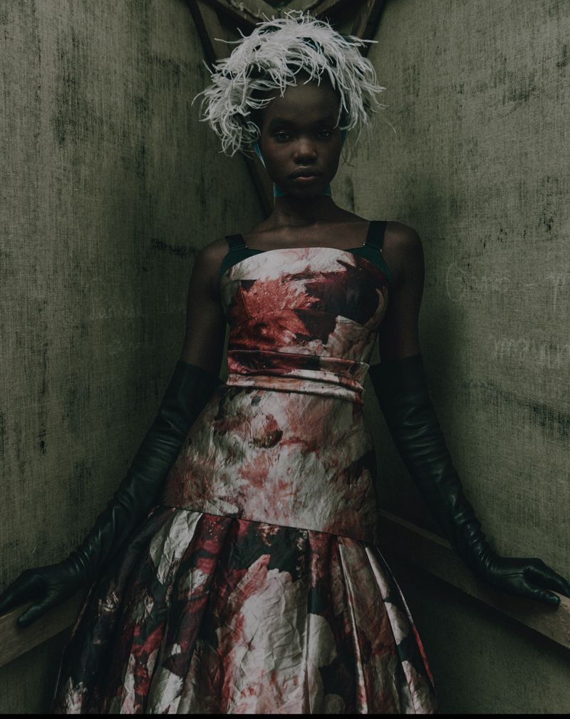 A woman in a floral dress and feathered headpiece stands in a dimly lit, textured corner, creating a dramatic and stylish fashion portrait.