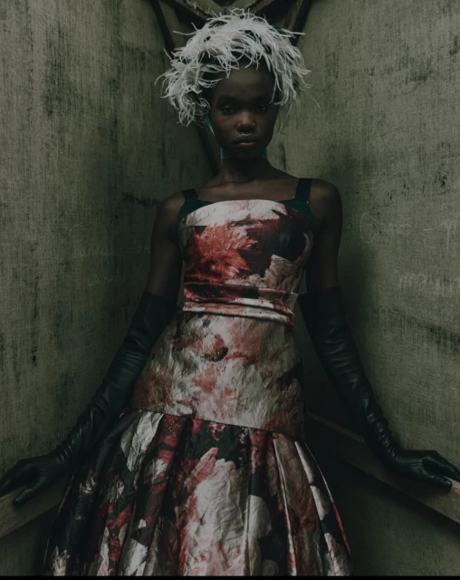 A woman in a floral dress and feathered headpiece stands in a dimly lit, textured corner, creating a dramatic and stylish fashion portrait.