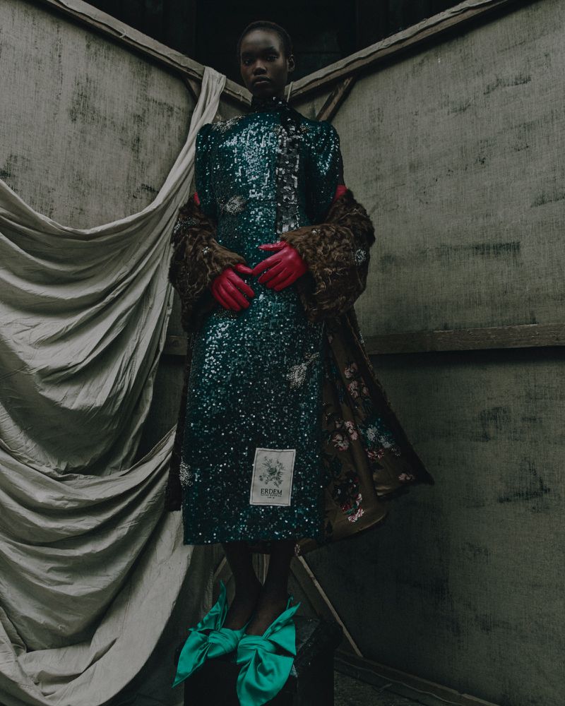 Fashion editorial shot of a Black female model in a green sequin dress and fur coat, posing indoors with draped fabric backdrop.