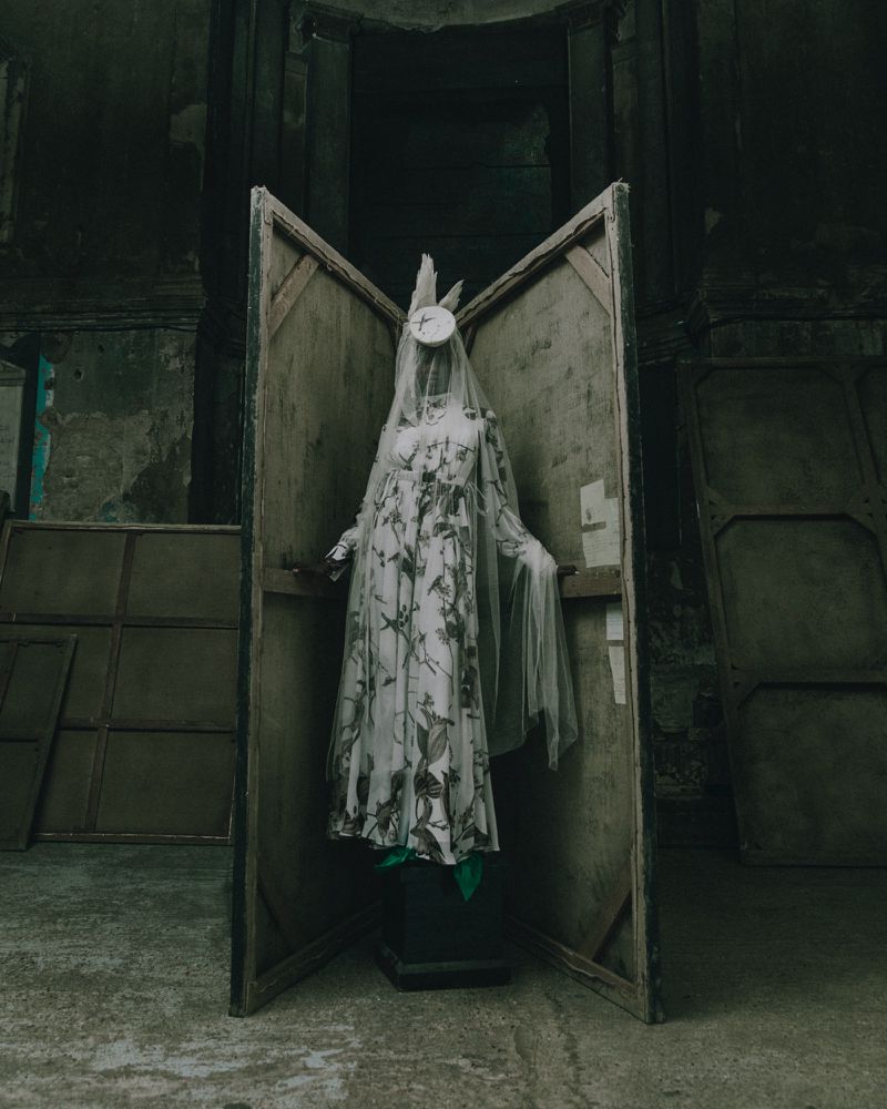 A veiled mannequin in a floral dress stands between wooden panels in a dim, dilapidated interior space, creating an eerie, artistic scene.