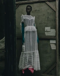Model Majda John Peter wearing a white lace dress with teal gloves standing against a large wooden canvas frame.