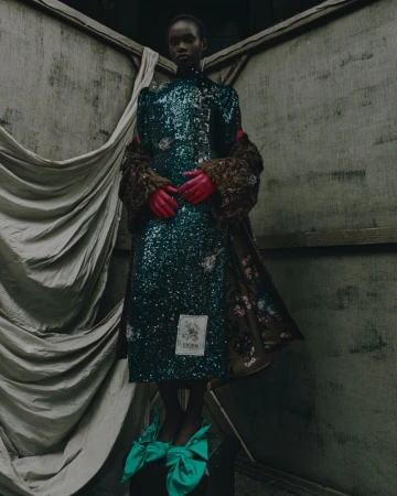 Model in a green sequin dress with red gloves standing against an industrial textured wall with draped fabric