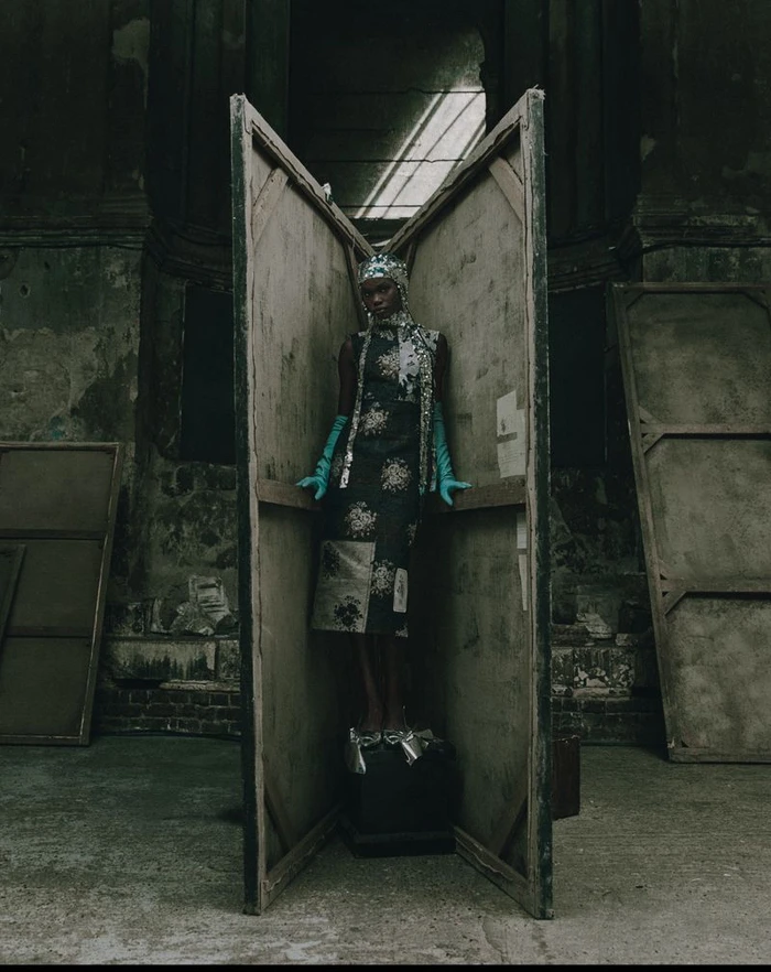 Model standing inside wooden folding screens wearing an embroidered dress and long teal gloves