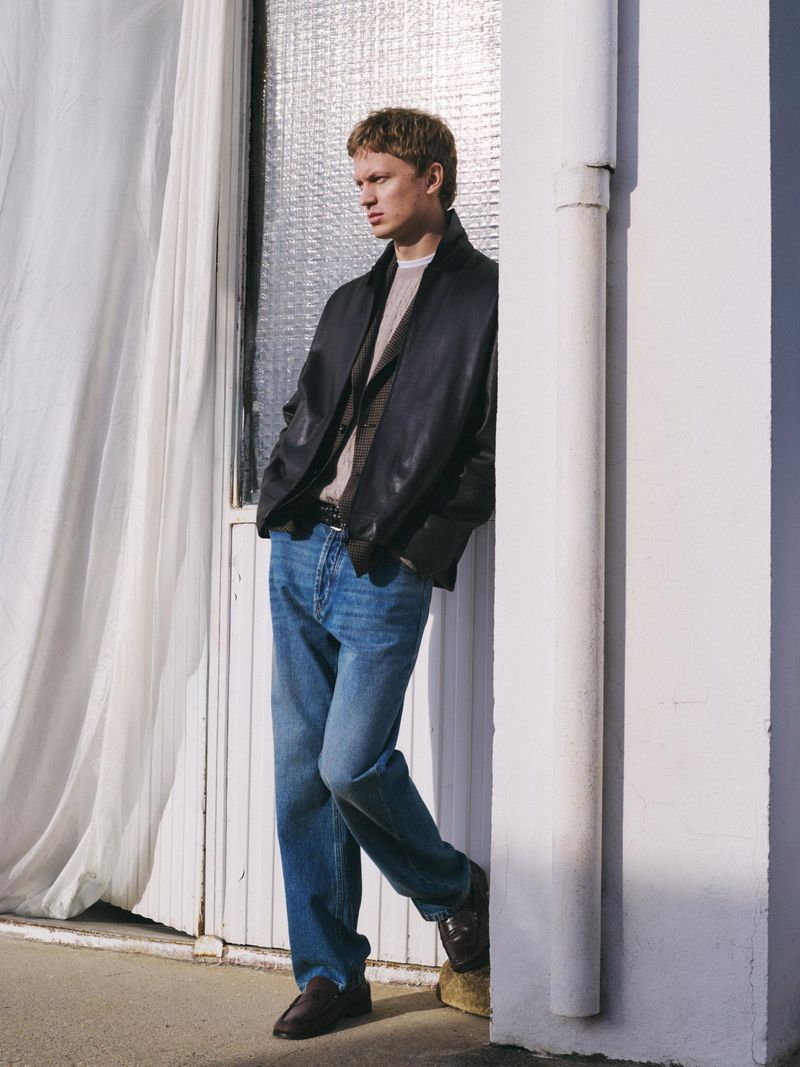 A blond man in a black jacket and blue jeans leans against a white wall next to a door with a window, showcasing a fashion-forward style.