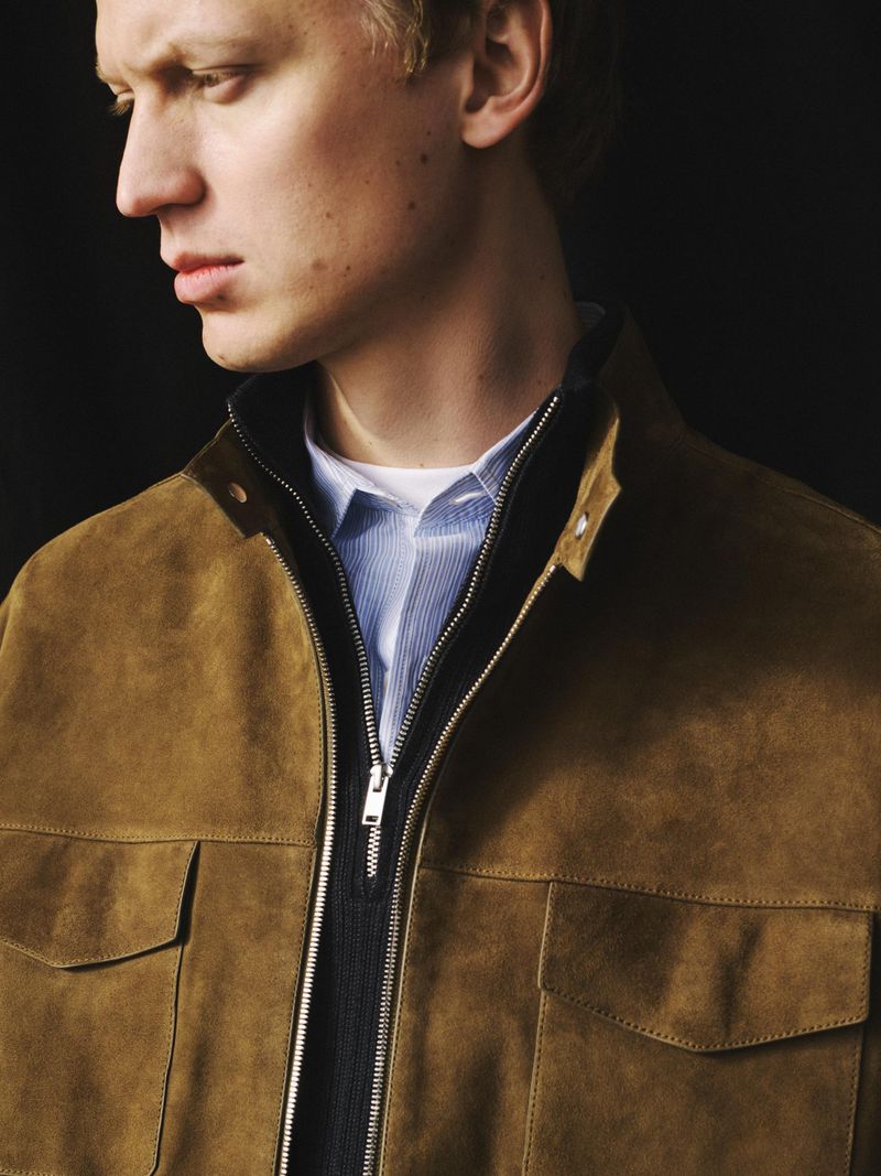 Close-up portrait of a male model wearing a camel suede jacket over a blue striped shirt and dark collar against a dark background.