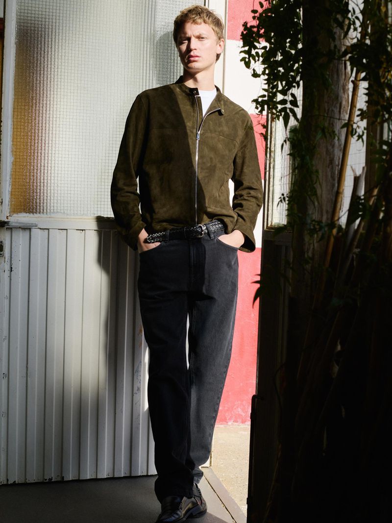 Full-length shot of a young adult male with blond hair posing outdoors in an olive-green jacket, gray denim jeans, and dark sneakers.
