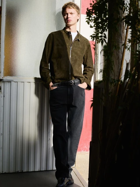 Full-length shot of a young adult male with blond hair posing outdoors in an olive-green jacket, gray denim jeans, and dark sneakers.