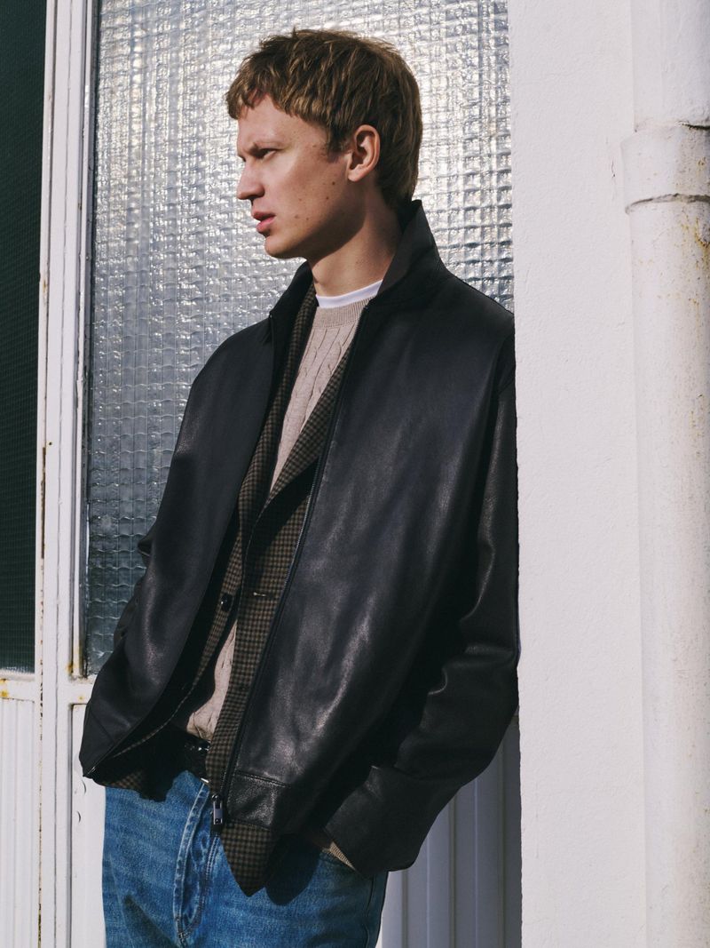 Man in black leather jacket, vest, and jeans leans against a white wall next to a window with a textured glass pane, looking left.