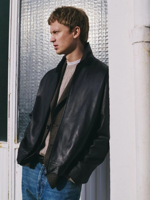 Man in black leather jacket, vest, and jeans leans against a white wall next to a window with a textured glass pane, looking left.