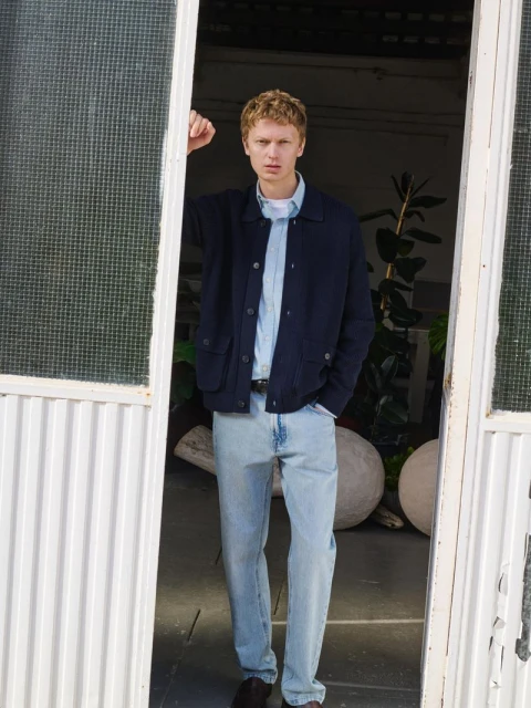 Full-body shot of a male model with blond hair wearing a navy jacket and light blue jeans, standing in a doorway with green plants.