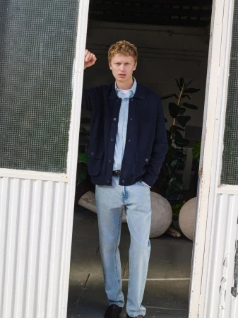 Full-body shot of a male model with blond hair wearing a navy jacket and light blue jeans, standing in a doorway with green plants.