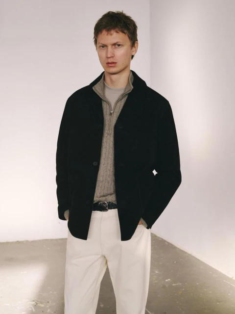 Adult male poses in a black jacket over a grey, quarter-zip sweater with white pants in a minimalist indoor photo studio setting.